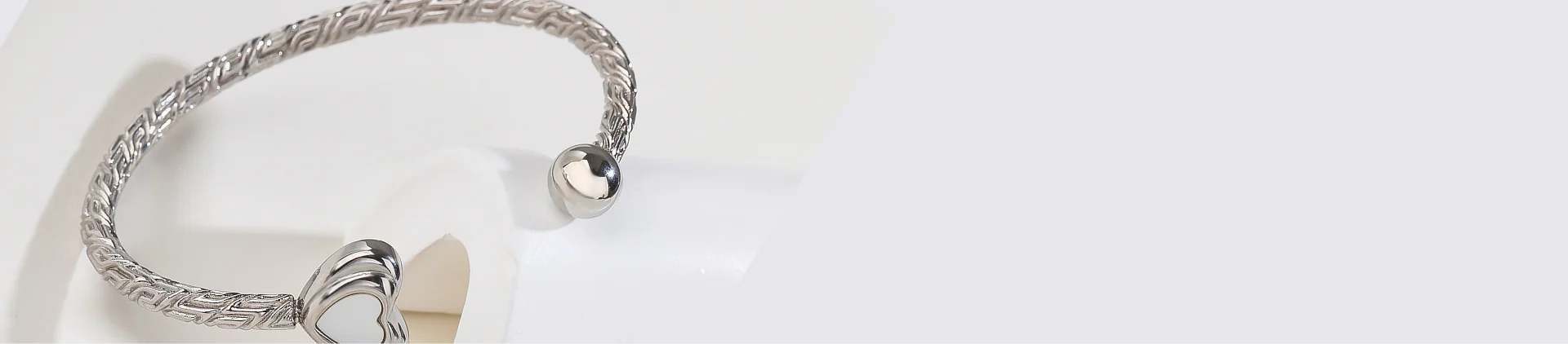 A silver-toned twisted wire cuff bracelet with a small heart charm, displayed on a white stand against a light gray background.
