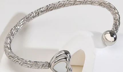 A silver-toned metal bangle bracelet with a braided Greek key pattern and a heart-shaped clasp, displayed on a white stand.