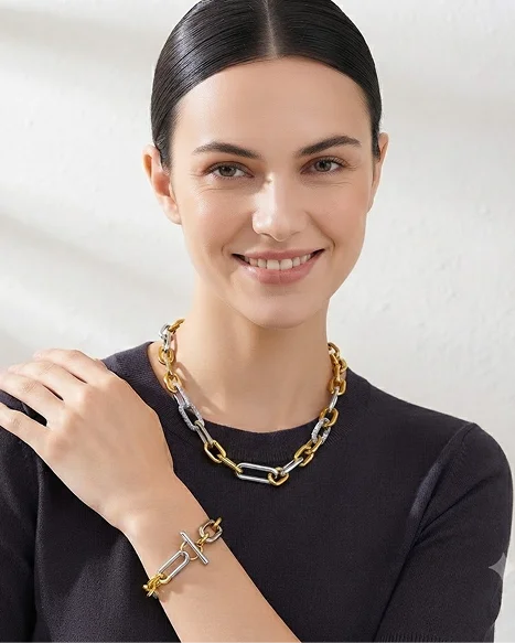 A woman with dark hair pulled back wears a gold and silver chain-link necklace and a matching gold and silver link bracelet, shown against a light textured wall.