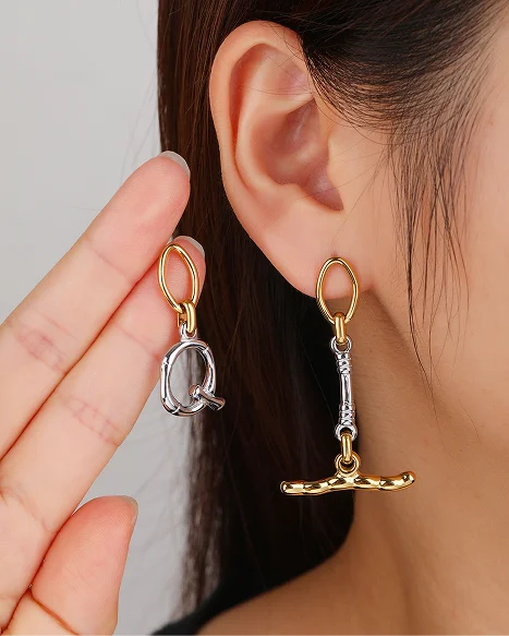 A close-up of a model's ear wearing a gold and silver dangling earring, with a hand holding the matching earring against a light gray background.
