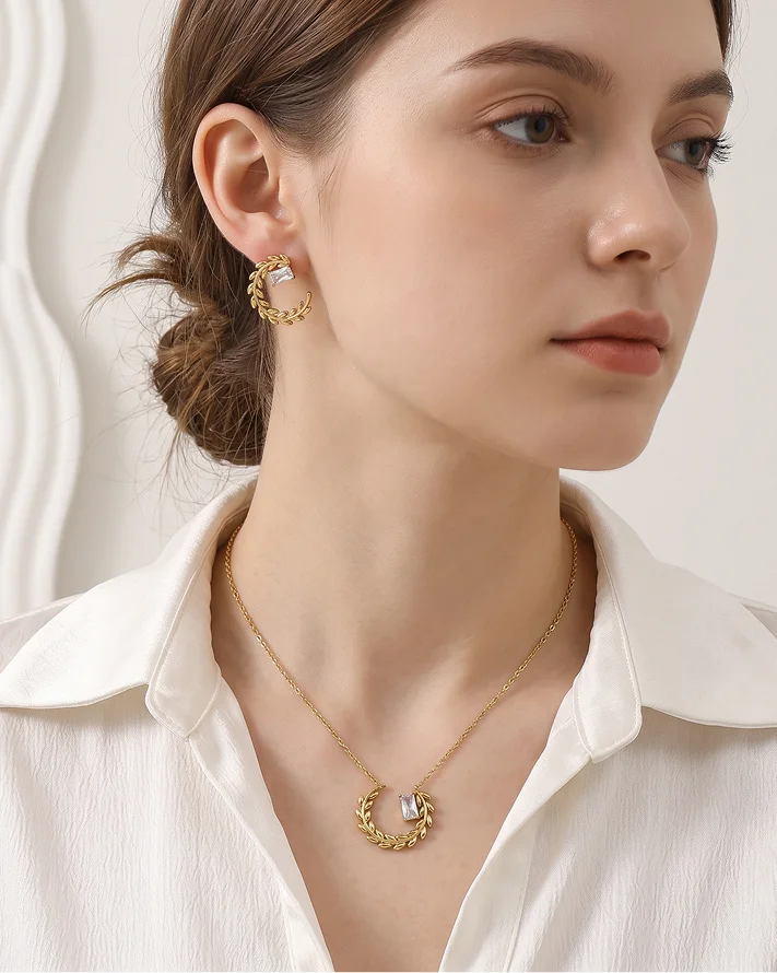 A woman wearing a delicate gold crescent moon necklace with a clear crystal center and matching gold hoop earrings, styled with a white collared blouse.