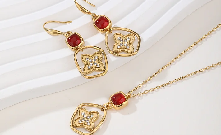 Gold jewelry set featuring a matching necklace and drop earrings with red gemstone accents and clear crystal details, displayed on a white wavy surface.